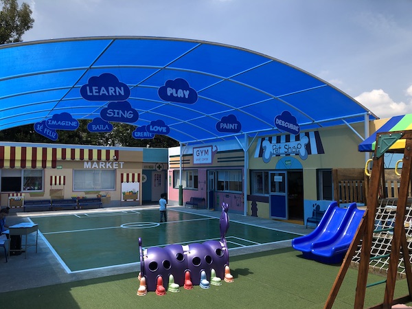 Patio de juegos exterior con techo y locales de pet-shop, gym y market alrededor.